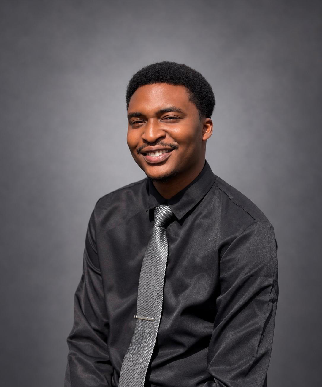 Pastor Pipeloluwa Oshinubi, smiling in a black shirt and silver tie, photographed against a grey studio backdrop